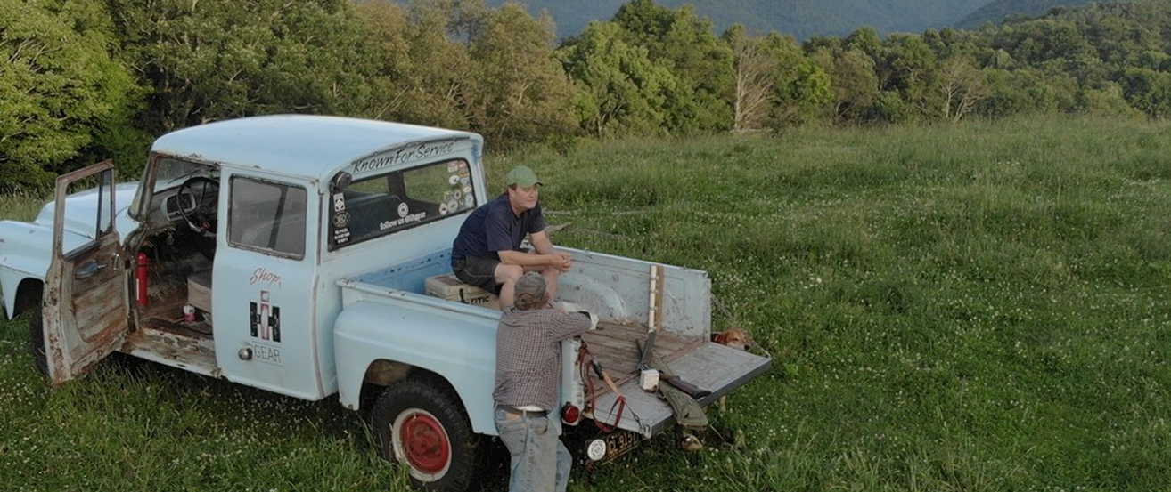 Two men working on a vintage truck in a grassy field with trees in the background